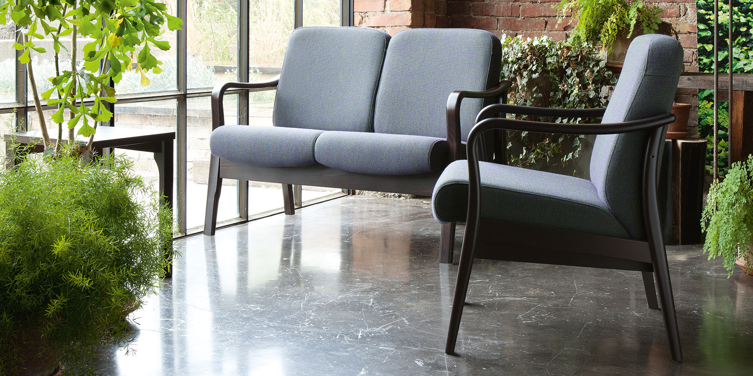 Gepolsterter Relax Sessel und zweisitziges Sofa mit Holzgestell in einem Raum mit Backsteinwand, großen Fenstern und Pflanzen